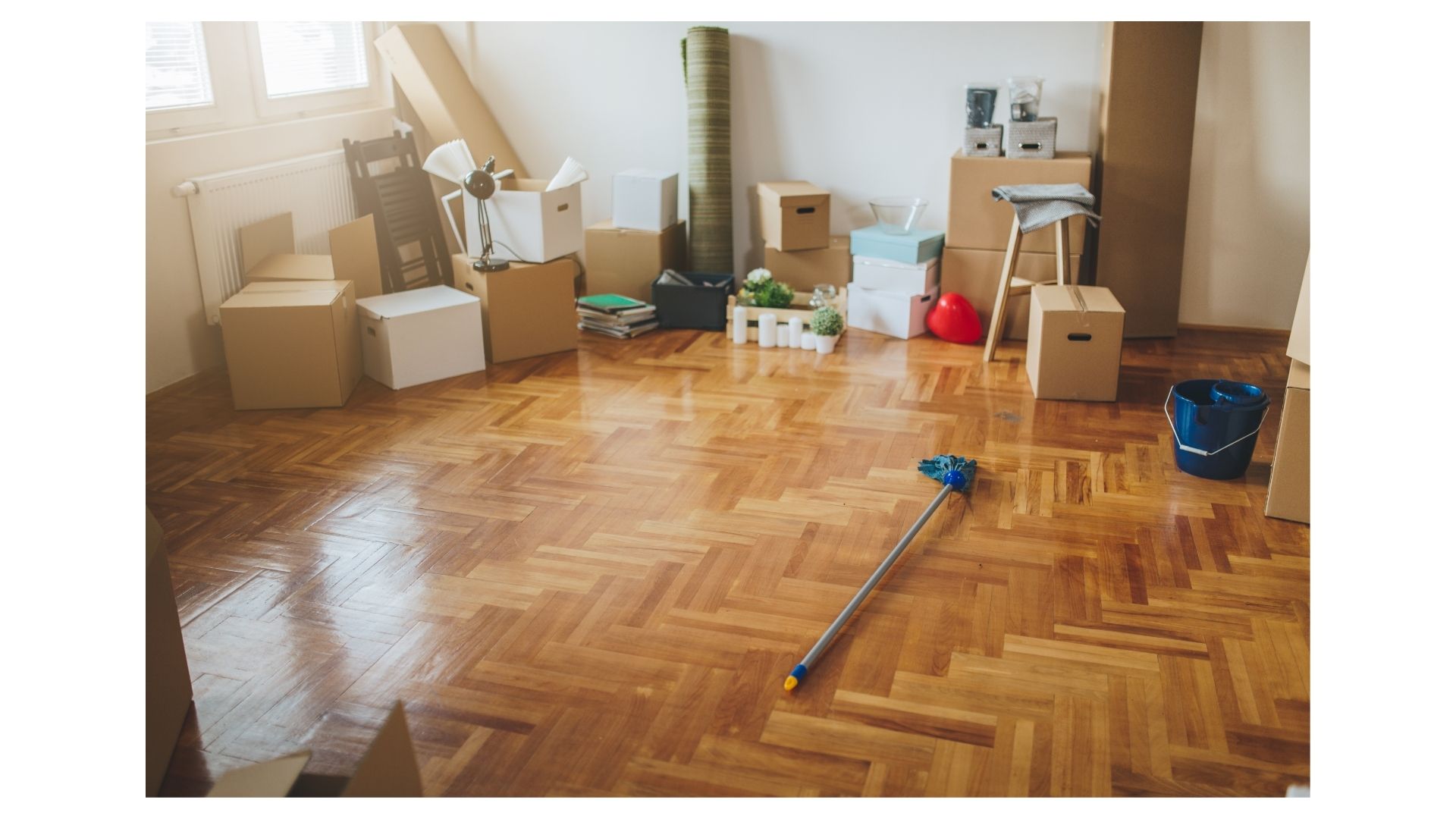 Room with packed cardboard moving boxes, cleaning supplies, and a mop on a polished wood floor