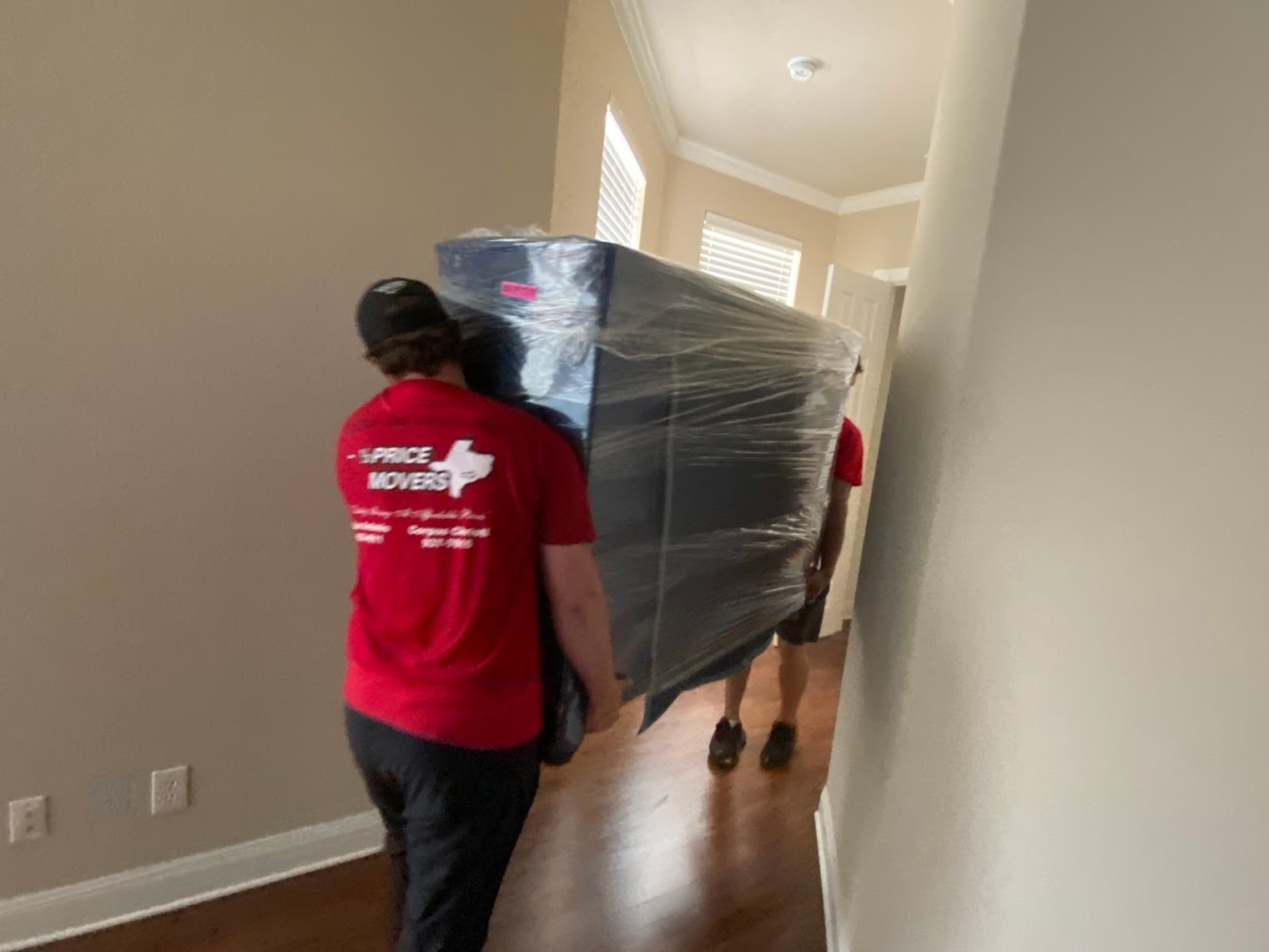 Two Half Price Movers crew members carrying a plastic-wrapped piece of furniture down an apartment hallway