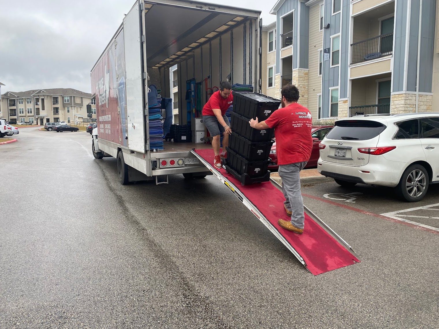 Two Half Price Movers crew members rolling a stack of moving bins up the ramp into a moving truck outside an apartment complex